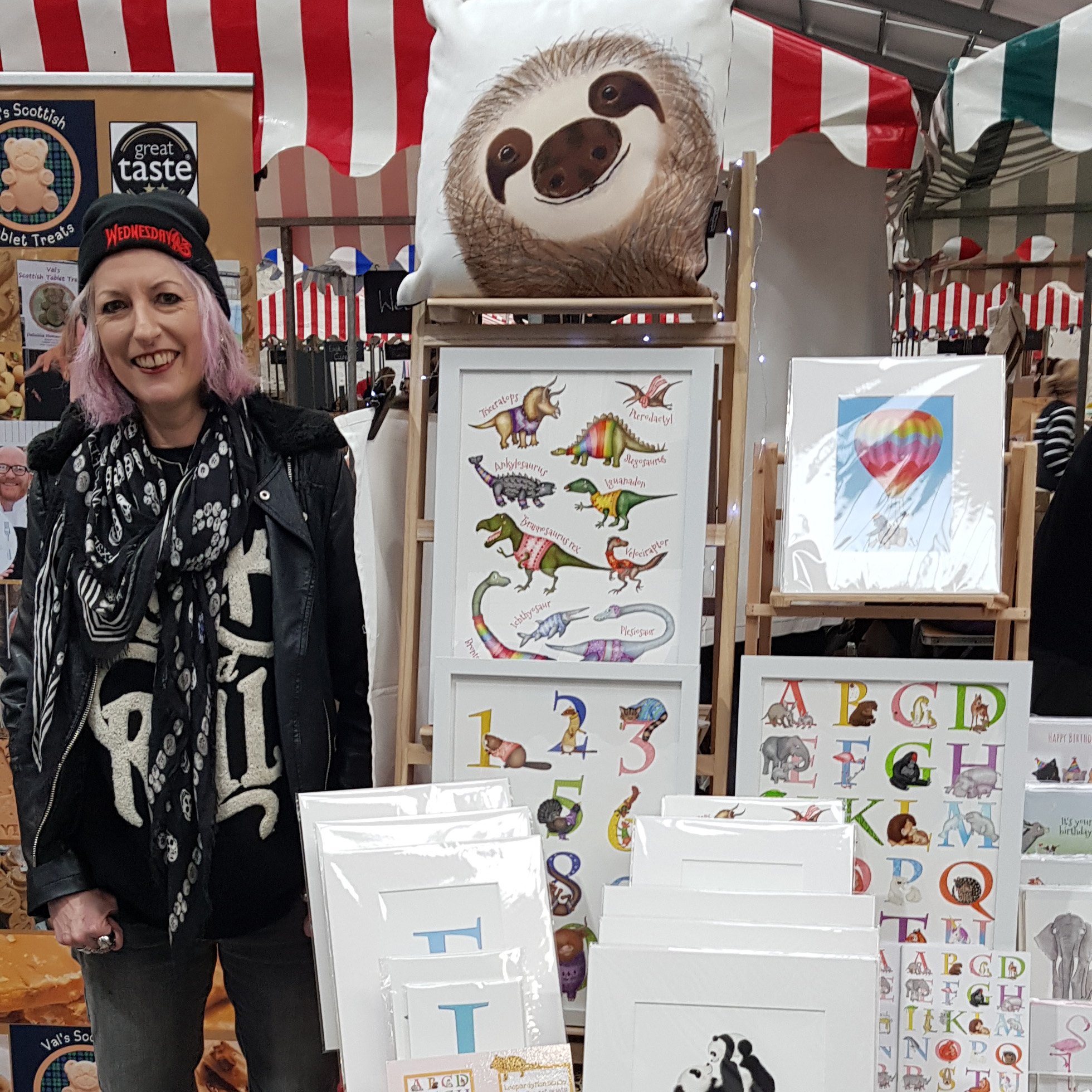 leopardynonsense-marketstall Elaine Macintyre, a white woman with pink hair wearing a beanie, standing beside the LeopardyNonsense market stall.