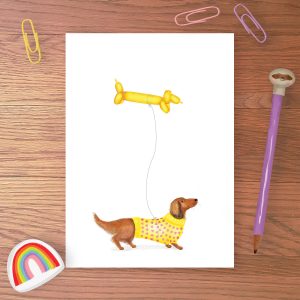 Greetings card with a cute dacshund wearing a yellow jumper and holding a sausage dog balloon on a string.