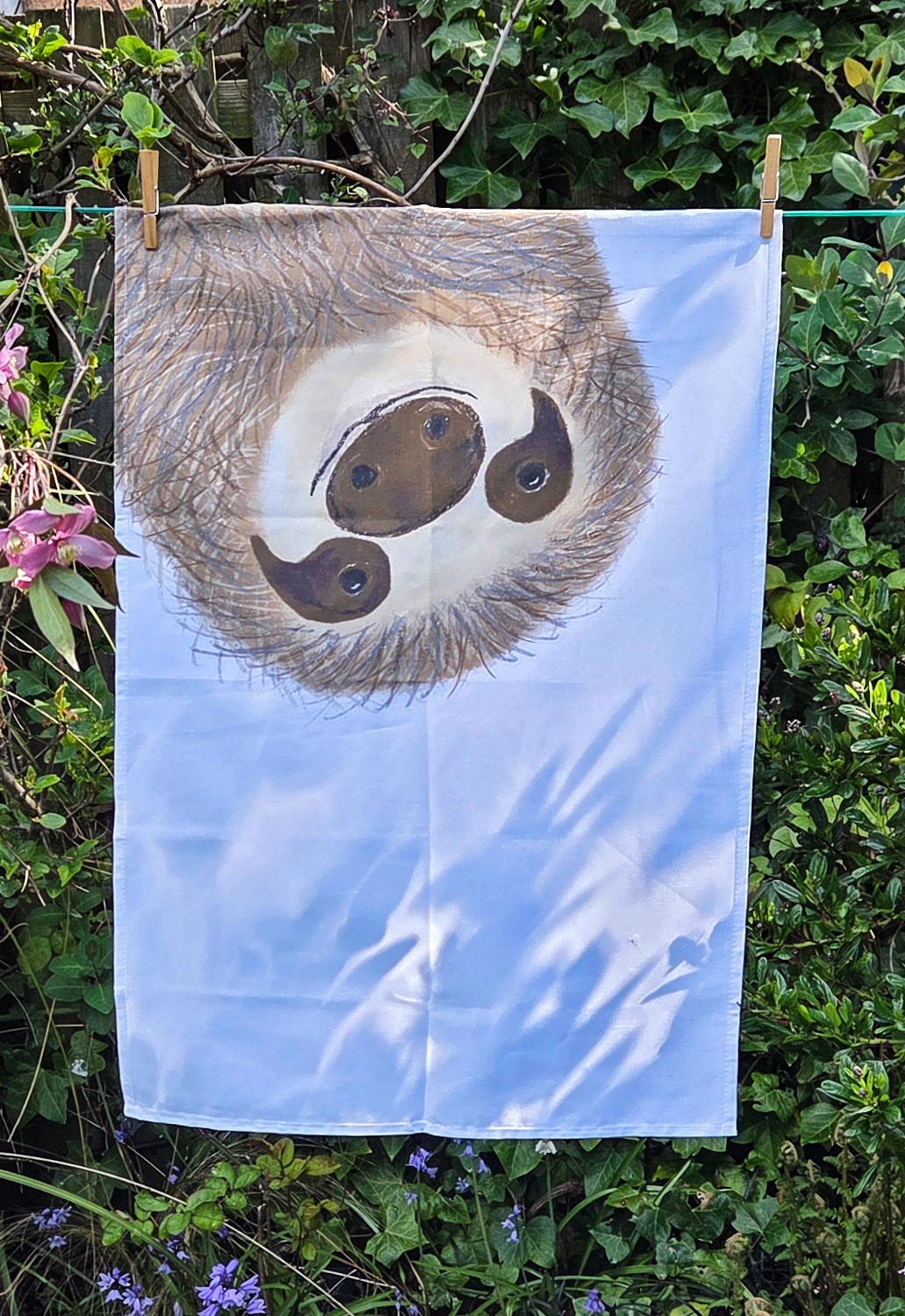Tea towel with a sloth's face on it, hanging on a washing line.