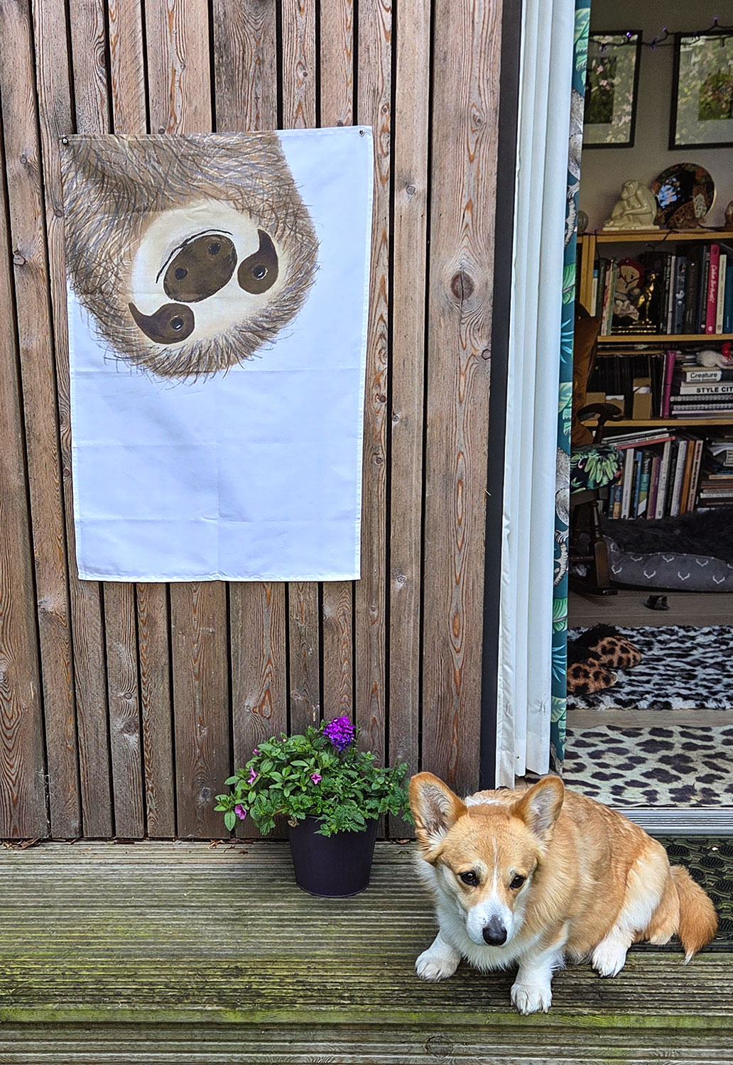 Tea towel with a drawing of a big smiling sloth's face, hanging up, with a corgi sitting beneath it.