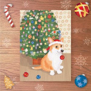 Christmas card with a cute corgi sitting in front of a Christmas treer, wearing a Santa hat and with a bauble in her mouth.